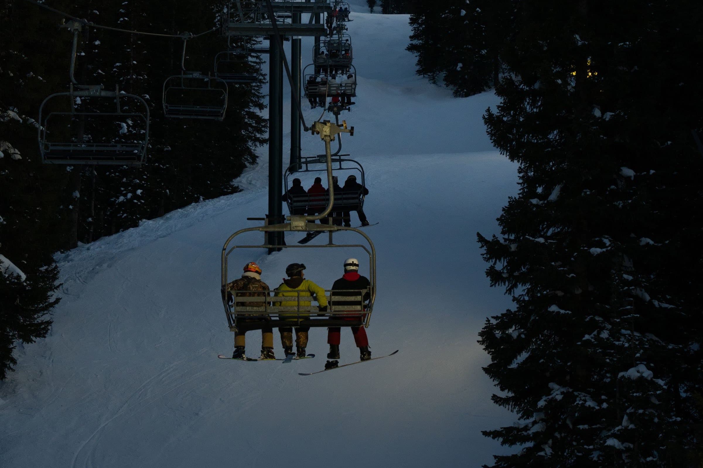 Three snowboarders riding a chairlift up a darkening run at dusk, snowy pines on either side.