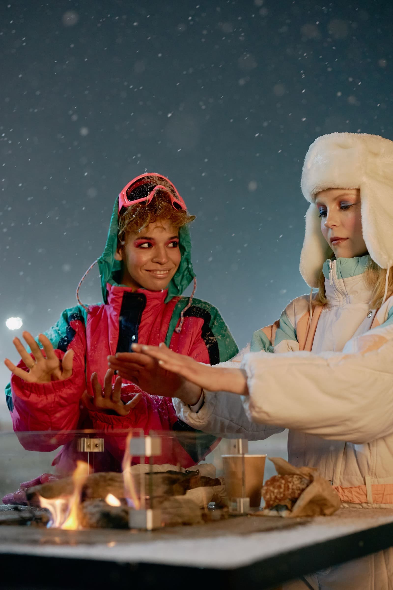 Two friends in retro ski suits warming their hands over a fire pit as snow falls at night.