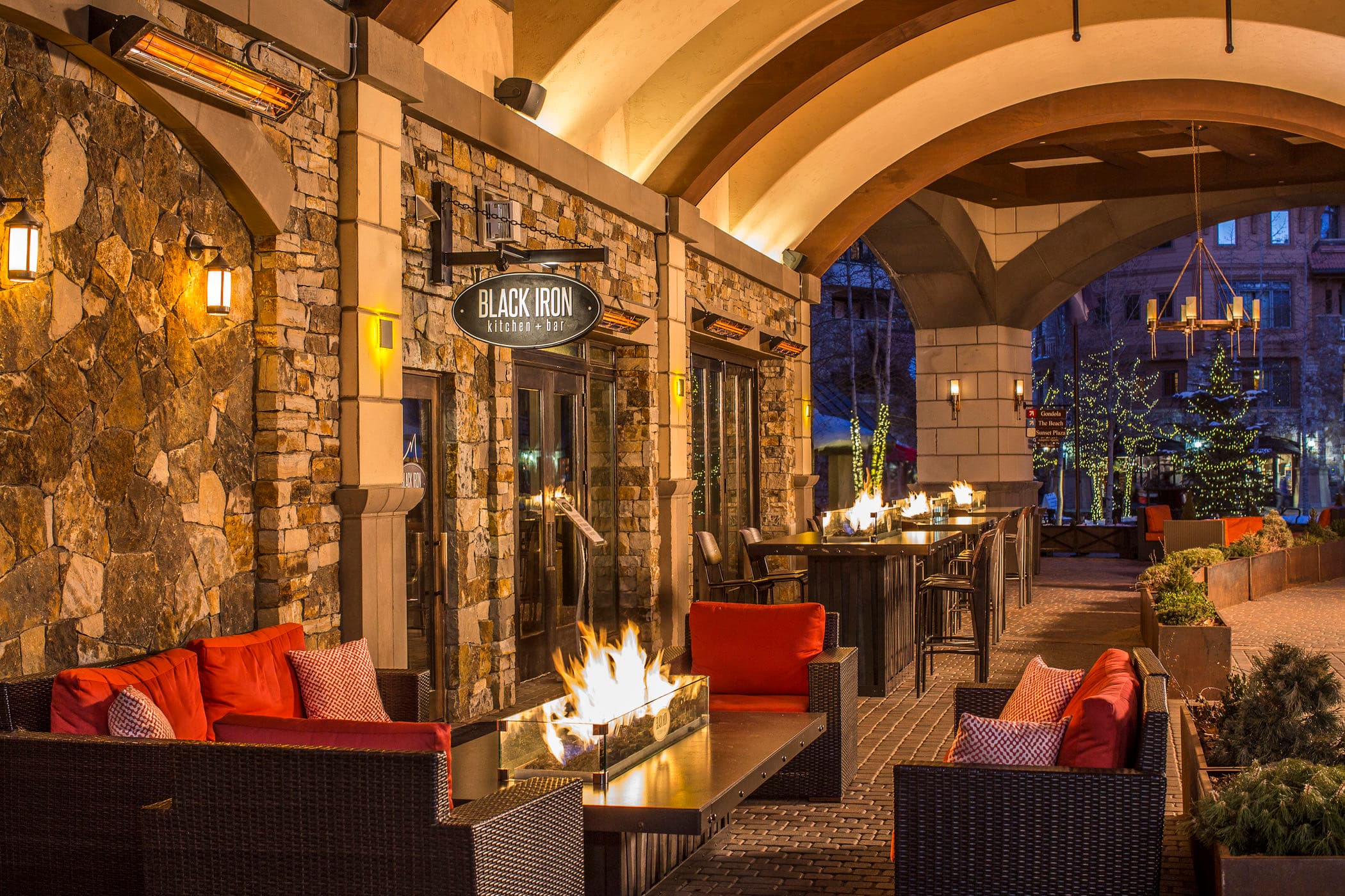 A stone terrace in Mountain Village at dusk, fire tables glowing under the arches and trees wrapped in lights.